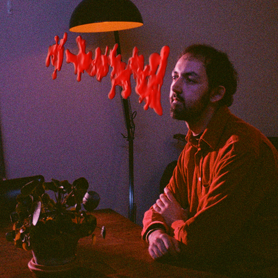 Lewis Coleman is sitting at a table in a dimly lit room with a lamp casting shadows. An abstract, blotchy red pattern hovers in front of Lewis' face.
